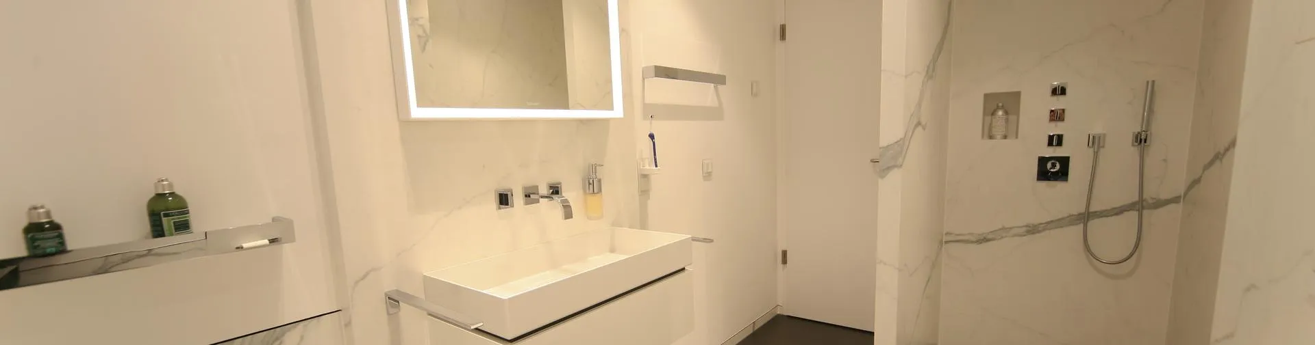 Modernes Badezimmer mit weißem Marmor, wandintegrierter Dusche, rechteckigem Waschbecken mit wandmontierter Armatur und beleuchtetem Spiegel.