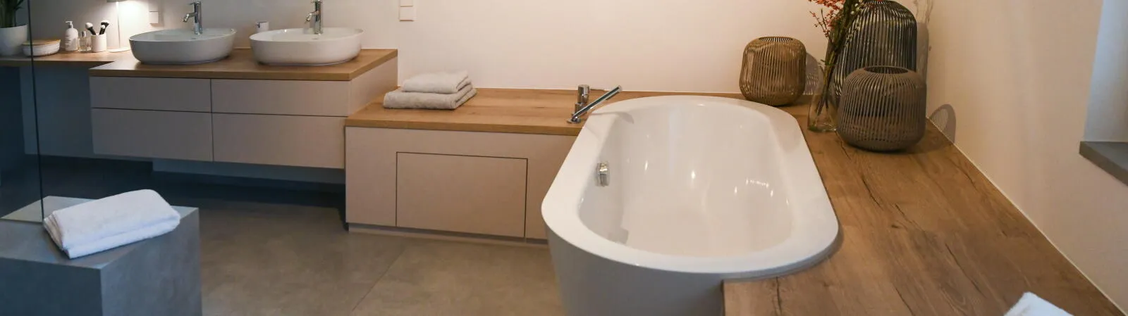 Modernes Badezimmer mit freistehender Badewanne, zwei runden Waschbecken auf einer Holzplatte und stilvoller Dekoration wie geflochtenen Vasen und frischen Handtüchern auf hölzerner Ablage.