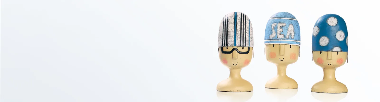 Drei dekorative Holzfiguren mit bunten Badekappen – gestreift, mit Schriftzug ‚SEA‘ und mit weißen Punkten – als Badeaccessoire-Dekoration.