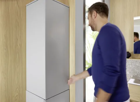 Ein Mann in einem blauen Pullover öffnet einen hohen, schlichten Schrank aus der burgbad Magictwist-Serie in einem Badezimmer.