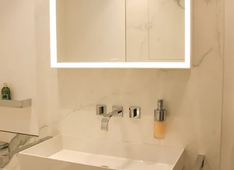 Detailansicht eines modernen Waschplatzes mit rechteckigem Waschbecken, wandmontierter Armatur, beleuchtetem Spiegel und Marmorwand im Hintergrund.