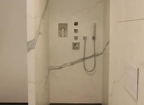 Begehbare Dusche mit Marmorverkleidung, integrierter Nische für Pflegeprodukte, moderner Handbrause und puristischer Armaturensteuerung in Chrom.