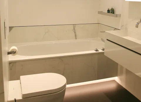 Modernes Badezimmer mit wandhängendem WC, rechteckiger Badewanne mit Marmorverkleidung, minimalistischem Waschbecken und indirekter LED-Beleuchtung am Boden.