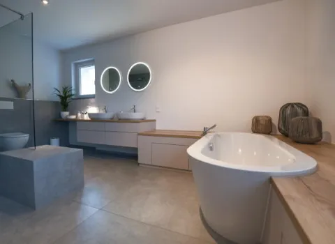 Geräumiges, modernes Badezimmer mit freistehender Badewanne, zwei Waschbecken unter runden, beleuchteten Spiegeln, einer großen begehbaren Dusche mit Glaswand sowie minimalistischer Dekoration auf Holzflächen.