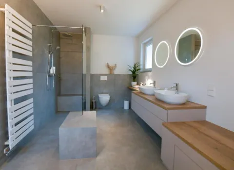 Modernes Badezimmer mit begehbarer Dusche, WC, zwei runden Waschbecken unter beleuchteten Spiegeln, großem Handtuchheizkörper und hellen Holz- sowie Naturstein-Elementen für ein minimalistisches Design.