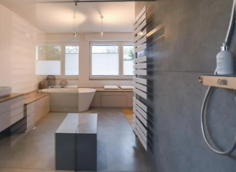 Blick durch eine Glasdusche in ein helles, modernes Badezimmer mit freistehender Badewanne vor drei Fenstern, Doppelwaschbecken, beheiztem Handtuchhalter und minimalistischer Einrichtung mit Holz- und Betonoptik.