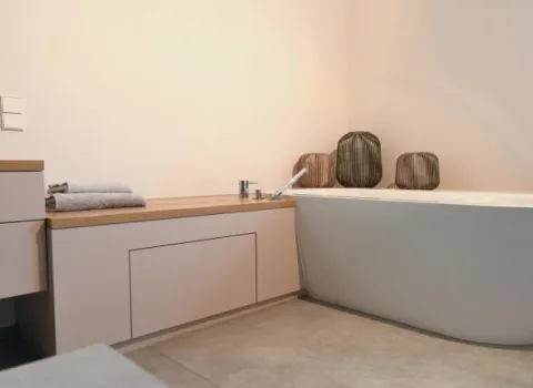 Detailansicht einer freistehenden Badewanne mit integrierter Armatur, daneben ein Holzboard mit gefalteten Handtüchern und dekorativen Vasen in einem modernen Badezimmer mit hellen, minimalistischen Möbeln.