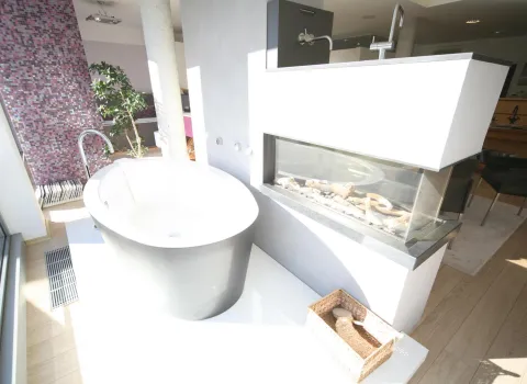 Moderne Badausstellung mit freistehender Badewanne neben einem Kamin mit Glasfront. Im Hintergrund eine Wand mit Mosaikfliesen in Lila- und Pinktönen sowie eine Grünpflanze als Dekoration.