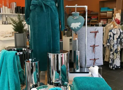 Auslage in einem Fachgeschäft für Bad- und Wohnaccessoires: Im Vordergrund türkisfarbene Handtücher, Badematte und Waschlappen, daneben glänzende Edelstahl-Badezimmerutensilien. Dahinter eine Schaufensterpuppe mit passendem türkisfarbenem Bademantel.