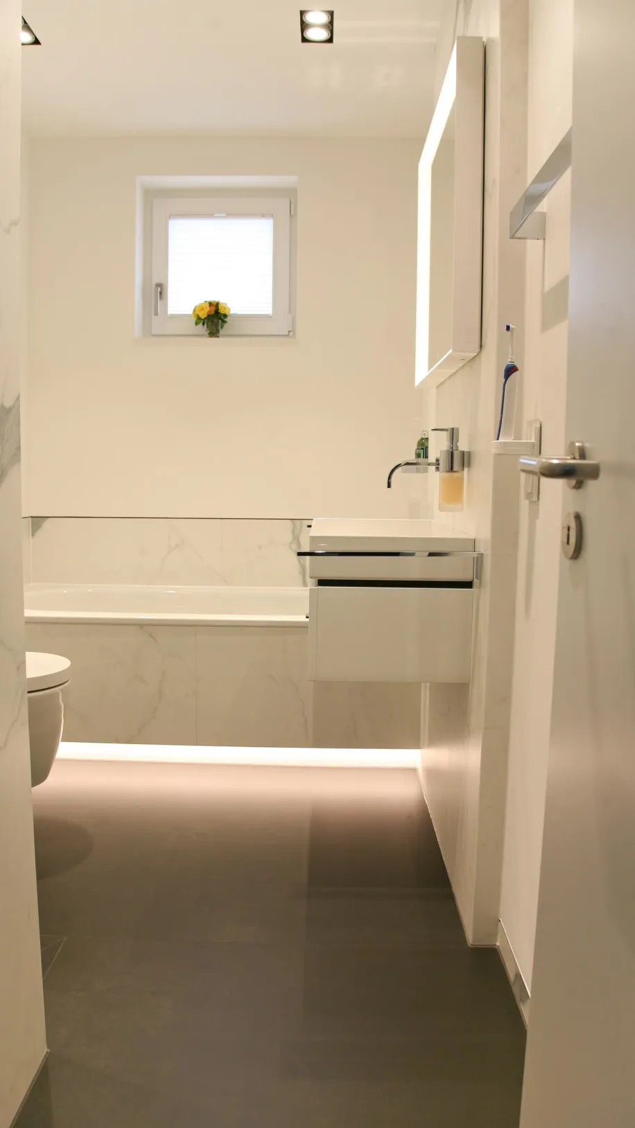 Helles, modernes Badezimmer mit freihängendem Waschbecken, wandhängendem WC, Badewanne mit Marmorverkleidung und LED-Beleuchtung am Boden sowie kleinem Fenster mit Blumenarrangement.