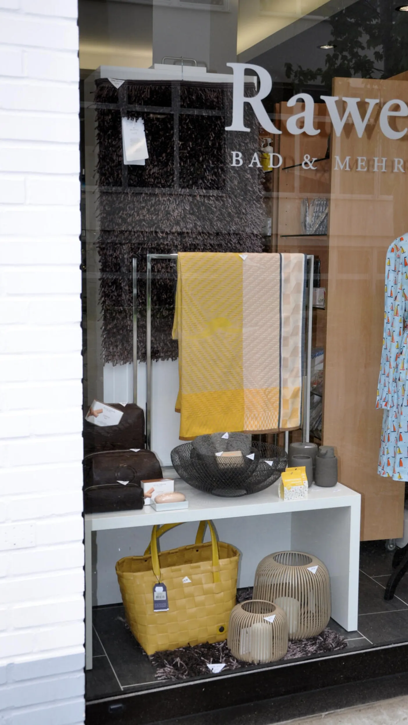Schaufenster von Rawe Bad & Mehr mit gelben und braunen Badezimmer-Accessoires: Handtücher in Gelb und Beige, Seifen und Schalen, dekorative Laternen sowie eine gelbe Einkaufstasche sind auf einem weißen Regal dekorativ arrangiert.