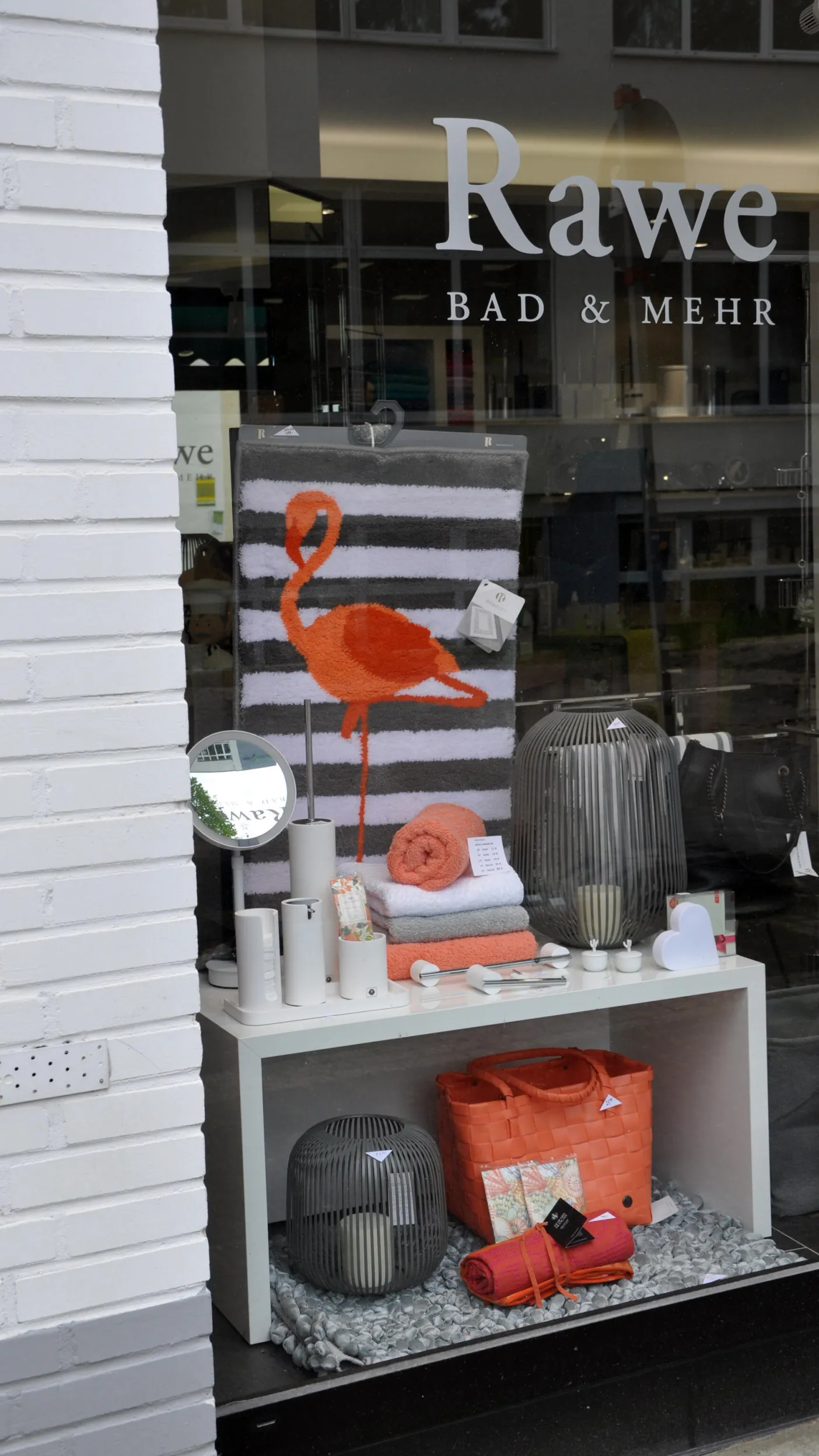 Schaufenster von Rawe Bad & Mehr mit dekorativen Badezimmer-Accessoires in Orange und Weiß: Auf einem Regal liegen Handtücher und eine Badematte mit Flamingo-Motiv, daneben Seifenspender, Spiegel und Laternen. Unten im Fenster sind ein orangefarbener Korb und weitere Dekoartikel ausgestellt.