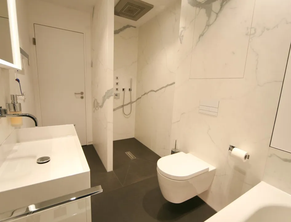Elegantes Badezimmer mit wandhängendem WC, bodengleicher Dusche mit Regendusche, modernem Waschbecken und großflächigen Marmorfliesen mit grauer Maserung.