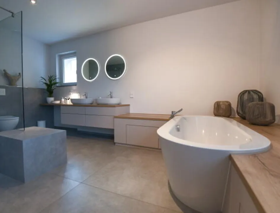 Geräumiges, modernes Badezimmer mit freistehender Badewanne, zwei Waschbecken unter runden, beleuchteten Spiegeln, einer großen begehbaren Dusche mit Glaswand sowie minimalistischer Dekoration auf Holzflächen.