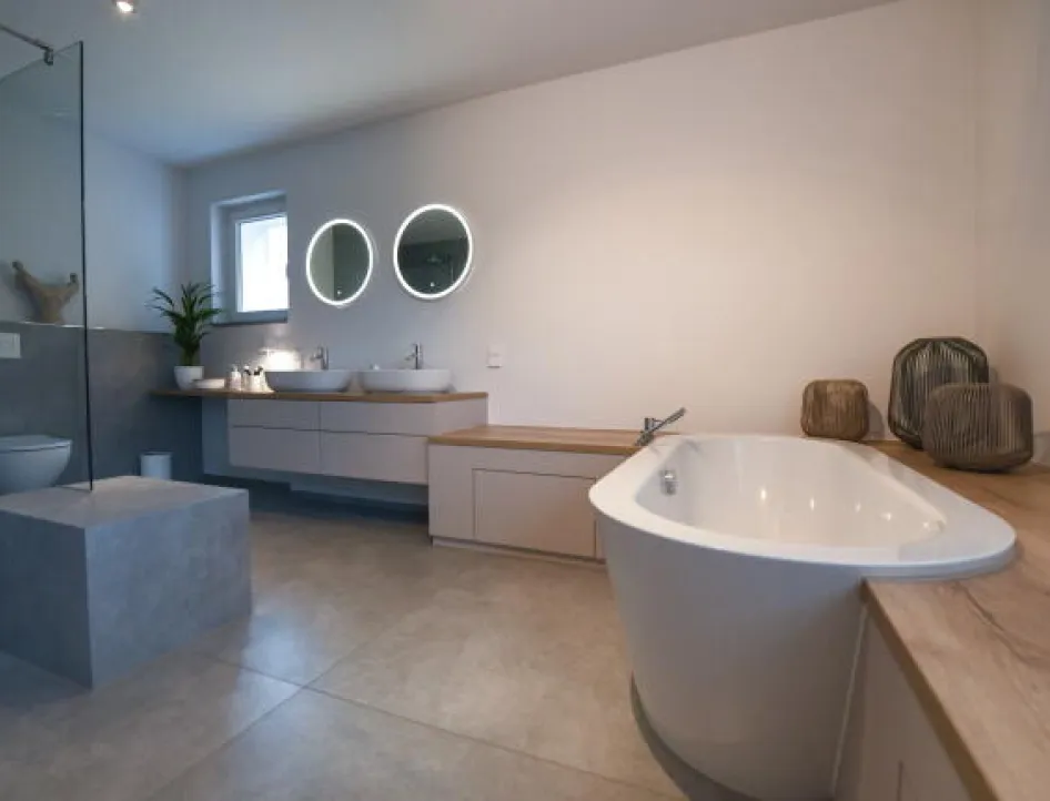 Geräumiges, modernes Badezimmer mit freistehender Badewanne, zwei Waschbecken unter runden, beleuchteten Spiegeln, einer großen begehbaren Dusche mit Glaswand sowie minimalistischer Dekoration auf Holzflächen.