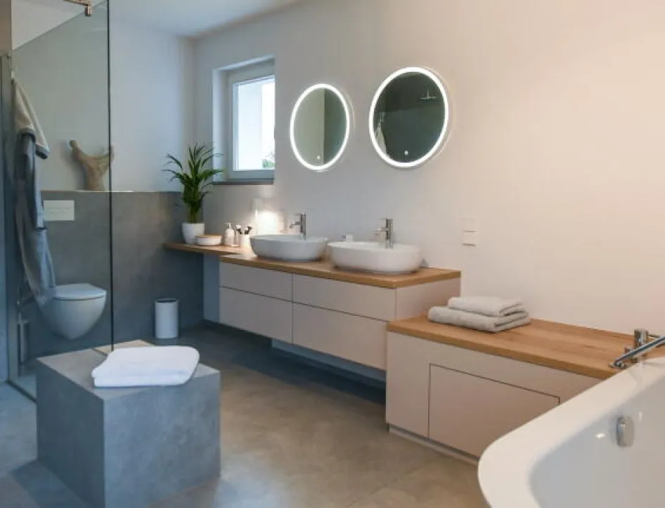 Modernes Badezimmer mit freistehender Badewanne, Doppelwaschtisch mit zwei runden Aufsatzbecken und beleuchteten Spiegeln, begehbarer Dusche mit Glaswand sowie hellen Holz- und Betonelementen.