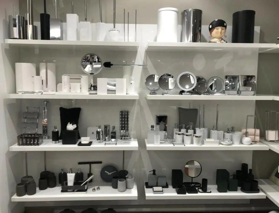 Regalwand in einem Geschäft mit modernen Badezimmer-Accessoires: Auf mehreren weißen Regalböden sind Kosmetikspiegel, Seifenspender, Zahnputzbecher, Toilettenbürstenhalter, Mülleimer und weitere Badutensilien in Weiß, Schwarz und Chrom ausgestellt.