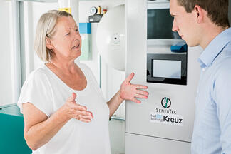 2 Personen im Beratungsgespräch vor Heizanlage Logo Kreuz